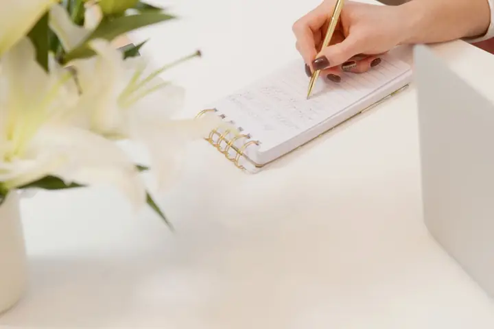 Beyaz masa üzerinde zambaklar ve not defterine yazan bir el
